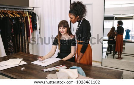 Fashion designer sketching a design sitting at her table while another woman looks on. Fashion entrepreneurs discussing a dress design in their fashion design studio.