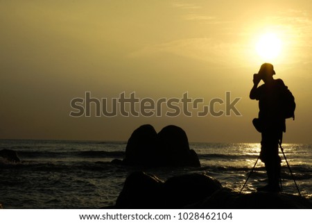 Silhouette of photographer at sunset on the beach using camera on tripod - Man taking picture in low brightness conditions standing by the ocean. Concept of travel, hobby and photography equipment