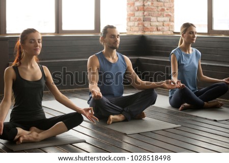 Group of three young sporty people practicing yoga lesson with instructor, sitting in Sukhasana exercise with eyes closed, Easy Seat pose, mudra gesture, working out, indoor close up, studio Royalty-Free Stock Photo #1028518498