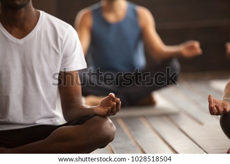 Young black man and a group of young sporty people practicing yoga lesson, sitting in Sukhasana exercise, Easy Seat pose, working out, indoor close up focus on mudra gesture, studio room Royalty-Free Stock Photo #1028518504