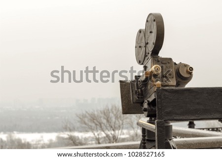 An old movie camera, against a sky background consisting of a tripod, lens, film coils.