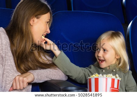 Pretty little happy girl feeding her mom with popcorn while enjoying  movie together at the cinema entertainment bonding childhood parenting love family happiness playful motherhood recreation.