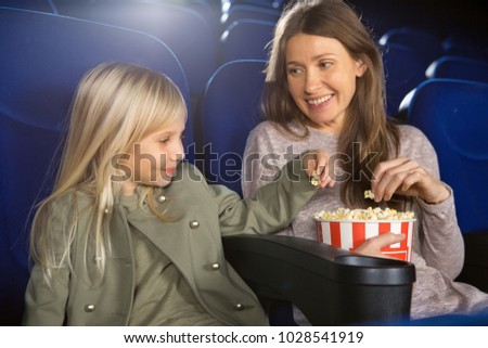 Adorable little girl eating ppcorn from the bucket her mother smiling happily while watching a movie together at the cinema cartoons children motherhood parenting recreation activity entertainment.