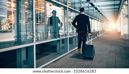 Rear view of young businessman walking outside public transport building with luggage. Business traveler pulling suitcase in modern airport terminal. Royalty-Free Stock Photo #1028616283