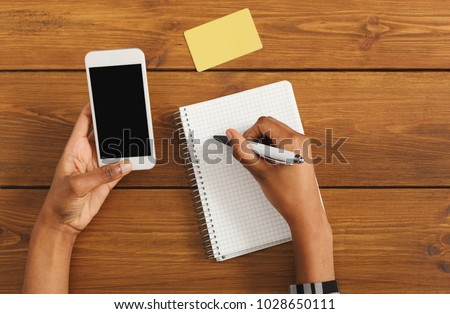 Black femael hands making notes in notebook and holding smartphone. Top view of african-american hands, laptop keyboard on a wooden table background. Education, business, technology concept