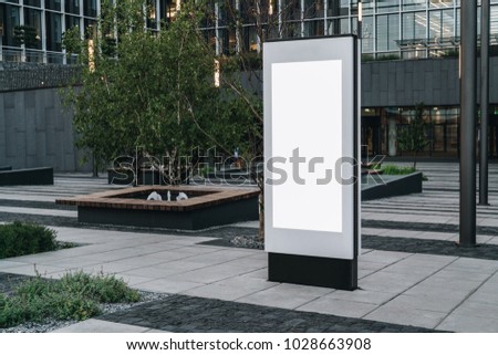 Summer evening. Vertical white blank billboard on city street. In background modern contemporary building. Mock up.