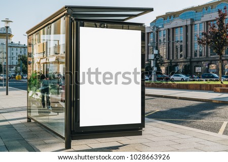 Vertical blank white billboard at bus stop on city street. In the background buildings and road. Mock up. Poster on street next to roadway. Sunny summer day. Royalty-Free Stock Photo #1028663926