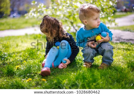 A boy and a girl are looking for Easter eggs among the grass. The brother and sister are sitting on the lawn with colorful eggs in their hands against the backdrop of flowering gardens.