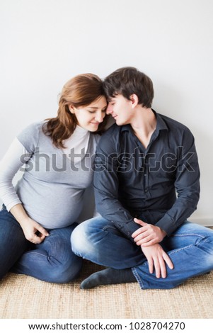 pregnancy, parenthood, fashion concept. on the floor by the white wall there is a man and a woman dressed in casual style, she is dressed in grey t shirt and he is in black shirt