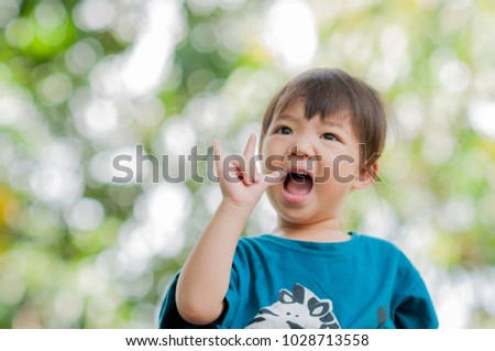 Lovely adorable asian girl laughing and making “i love you” hand sign with green bokeh background. Cheerful cute Asia kid (three years old) showing “i love u” symbol with hand in the blurred garden.
