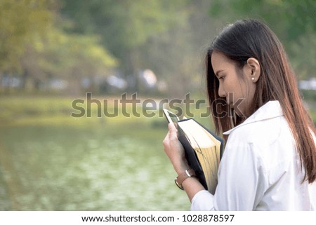 Beautiful asian woman using smart phone outdoors and holding a book at garden park in the evening with sunset time.