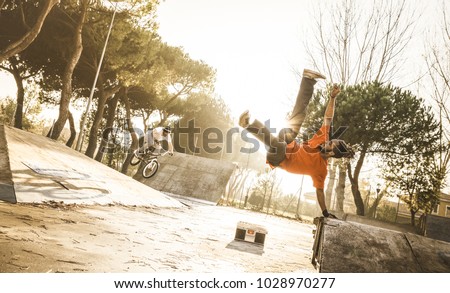 Urban athlete breakdancer performing acrobatic jump flip at skate park - Guy riding bmx bicycle behind mate acrobat dancing with extreme move - Breakdance and friendship concept - Warm sunset filter Royalty-Free Stock Photo #1028970277