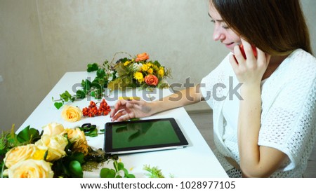 Alluring girl florist communicates with  supplier of colors on  smartphone, smiles and makes appointment, checks information about  order and drives  fingers along  touch screen of  tablet. Wo
