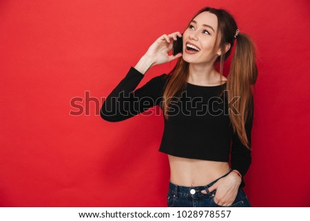 Photo of young happy lady standing isolated over red background talking by mobile phone. Looking aside.
