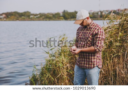 Young unshaven man in checkered shirt and cap pulls out maggots bait from small box to put her on rod against background of lake, shrubs and reeds. Lifestyle, fisherman recreation, leisure concept
