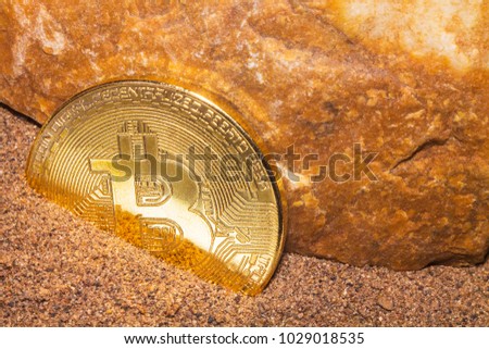 The gold coin of the crypto currency Bitcoin lies in the sand near the orange rock.