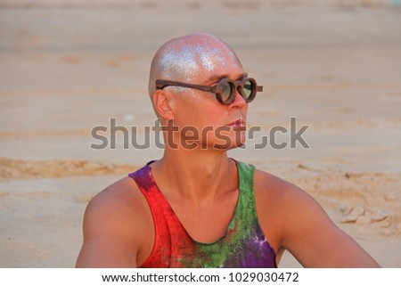 Bald man freak in bright clothes and round glasses at a freak parade or festival, on the beach. India, Goa. An unusual man, the image of a designer's man.