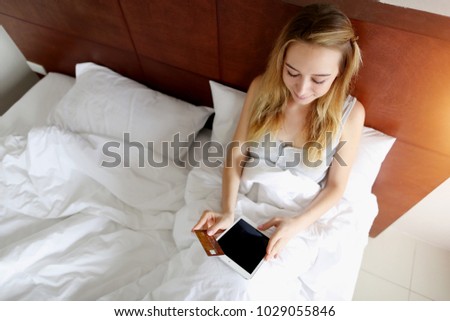 woman transfer money holds debit credit card and tablet in white bed at home. concept of new technologies, online shopping buy goods with sunshine