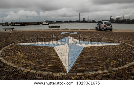 A view of an empty embankment on a cloudy day, IJmuiden,the Netherlands. There is a wind rose pointing to the North,two benches,a car on the boardwalk,a ship sails by sea and a steelworks on the photo
