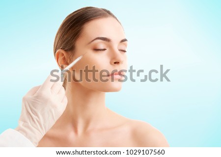 Portrait of an attractive young woman receiving botox treatment. Isolated on light blue background. Royalty-Free Stock Photo #1029107560