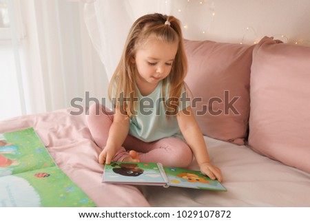 Cute little girl reading book while sitting on bed at home