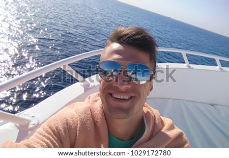Young handsome smiling man in sunglasses making selfie or photo on the phone riding on a yacht at sea.