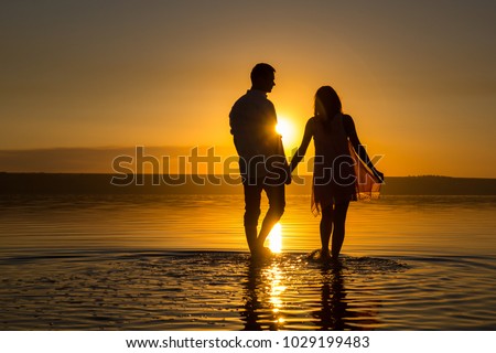 Young couple is walking in the water on summer beach. Sunset over the sea.Two silhouettes against the sun. Just married couple hugging. Romantic love story. Man and woman in holiday honeymoon trip.
