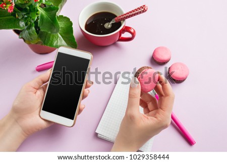 Female hands holding phone. Top view on french macarons on trendy color pink desk. Woman and stilish workplace. Cup of coffee, phone, notebook. Breakfast, cakes, coffee. Women's Day concept
