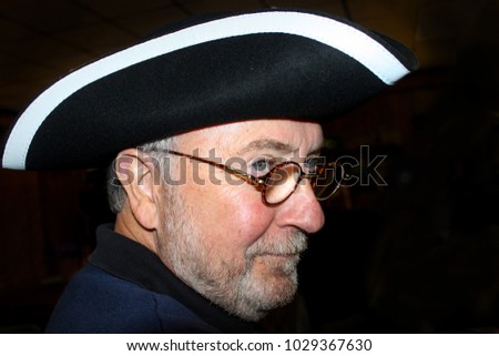 Greyhaired man with tricorn hat and round glasses and sly smile