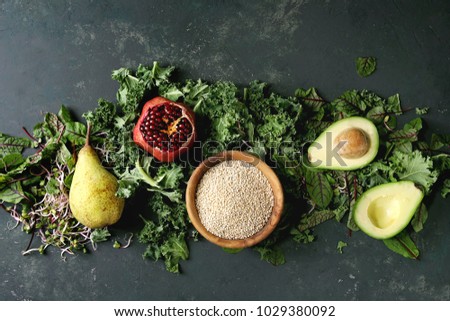 Green salad and fruits mix for salad with kale, young beetroot leaves, sprouts, pear, avocado, pomegranate, bowl of raw uncooked quinoa over dark texture surface. Top view, space. Food background.