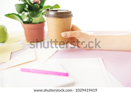 Female hands holding coffee. Side view on trendy color pink desk. Woman and stilish workplace. Cup of coffee, phone, notebook. Breakfast, coffee. Women's Day concept. Flowers