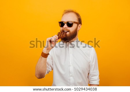 Cheerful young bearded hipster man eating a bar of chocolate over orange background. Unhealthy concept. Royalty-Free Stock Photo #1029434038