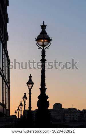 Row of tall elegant old lit street lamps silhouetted against the sunset sky at dusk along the London South Bank
