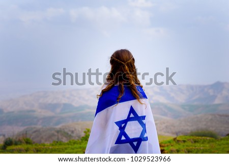 Little patriot jewish girl standing and  enjoying great view on the sky, spring field and mountains with the flag of Israel wrapped around her. Memorial day-Yom Hazikaron and Yom Ha'atzmaut concept.