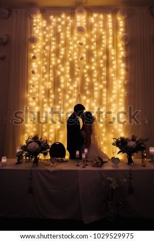 The kiss of the bride and groom. Wedding banquet in the hall decorated with light bulbs. Presidium. Celebration. Table setting. Bride and groom. Kiss. Silhouette of a couple.