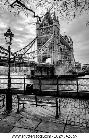 A black and white photograph of Tower Bridge in London with a park bench and cobblestone road in the forground