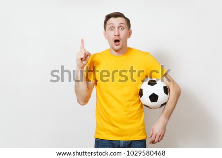 Young shocked European man, fan or player in yellow uniform hold soccer ball, point index finger up, cheer favorite football team isolated on white background. Sport play football, lifestyle concept