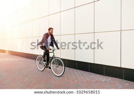 Full length photo of smiling young bearded man riding a bike on the city street. Dressed up in plaid shirt, t-shirt and jeans. Recreation concept.