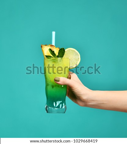 Blue and green tropical alcohol cocktail drink with pineapple in woman hand on mint background