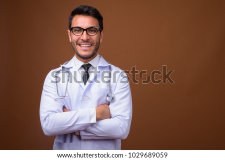 Studio shot of young handsome Hispanic man doctor wearing eyeglasses against brown background