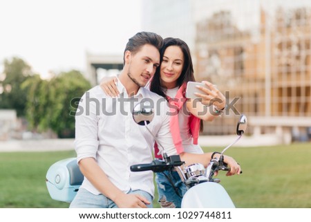 Refined girl with black hair and cute smile making selfie with boyfriend on date in summer weekend. Blissful young couple in stylish clothes sitting on scooter and taking picture of themselves.