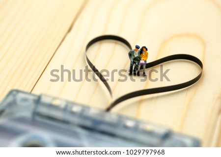 Miniature people : Couple of love sitting on Audio cassette with magnetic tape in shape of heart on wood background.