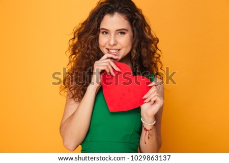 Picture of beautiful young lady standing isolated over yellow background. Holding heart looking camera.