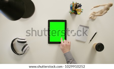 A finger touching a green screen of a tablet. The tablet is on the white table. View from the top. Close-up.
