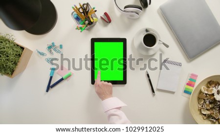 A finger touching a tablet with a green screen. The tablet is on the white table. View from the top. Close-up.