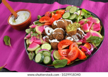 tasty falafel balls with greek yogurt sauce on plate with vegetable colorful salad of watermelon radish, chard leaves, cucumber and tomato slices. tzatziki sauce in bowl, view from above, close-up