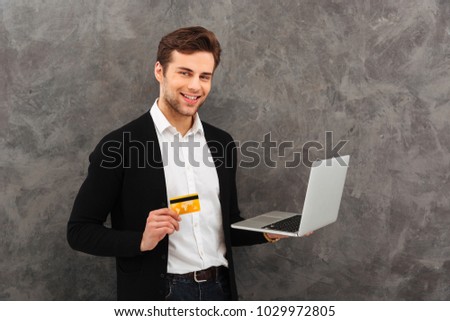 Photo of young happy businessman standing over grey wall background. Looking camera using laptop computer holding credit card.