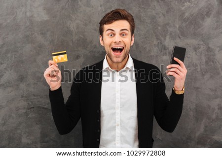 Picture of young excited businessman standing over grey wall background. Looking camera chatting by mobile phone holding credit card.
