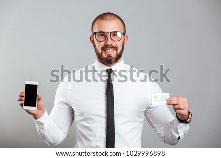 Photo of happy successful man in glasses and tie holding cell phone and credit card isolated over gray background