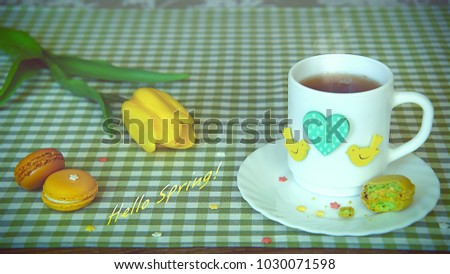 Shot with lettering "Hello Spring!", using yellow tulip, a white cup of tea decorated with green heart and yellow birds, and colorful macaroons, cookies, checkered tablecloth, medium shot